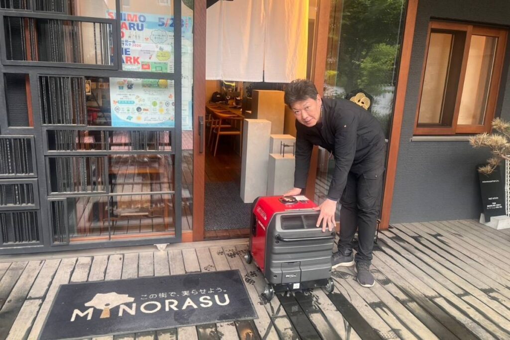東京都大田区にあるミノラスパートナーズ株式会社の正面玄関の前で発電機を持っている亀山くん